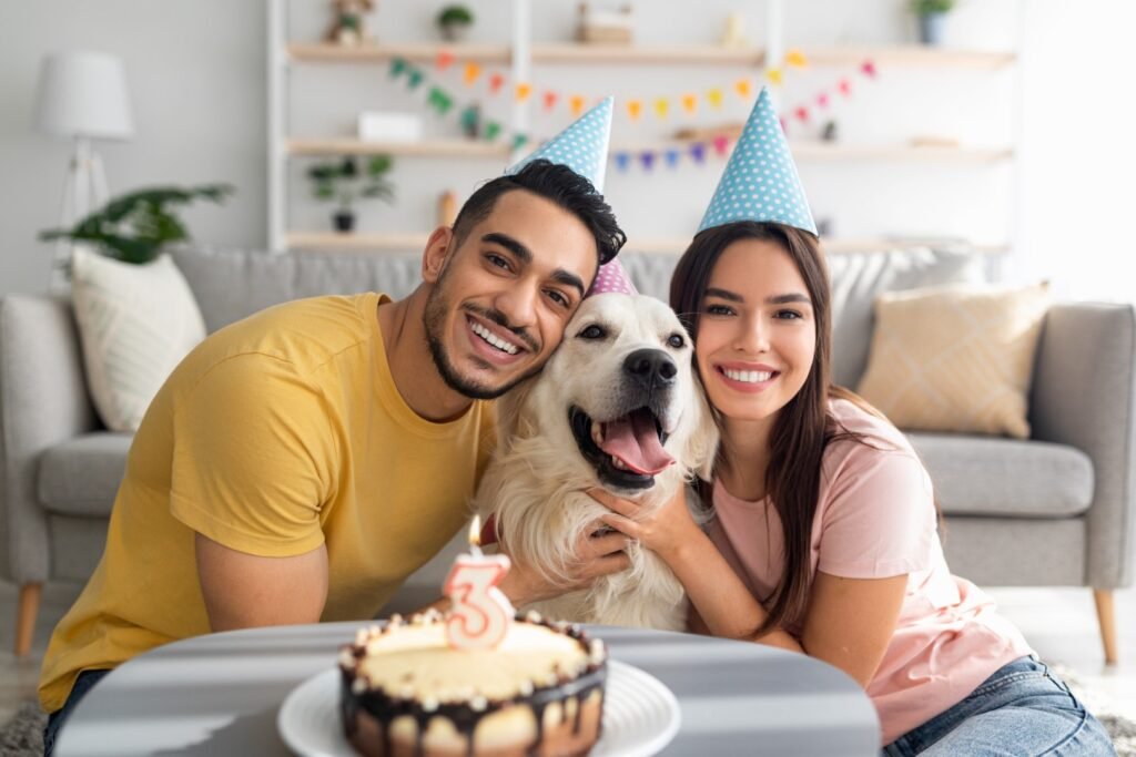 Happy multiracial couple celebrating their dog's birthday with festive cake, wearing party hats at home. Cute golden retriever having b-day with his owners, enjoying anniversary
